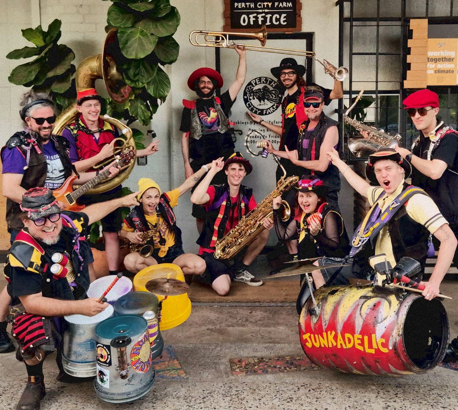A group of smiling people holding percussion and brass instruments pose in front of a white building